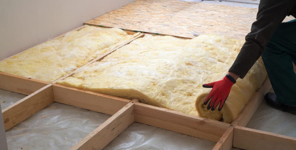 Personne installant de la laine de verre jaune dans un cadre de bois pour l'isolation d'un plancher, avec des panneaux OSB posés.