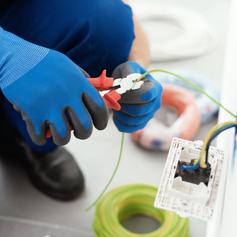 Électricien ganté coupant un fil électrique vert avec une pince. Prise murale et rouleaux de câble en fond.