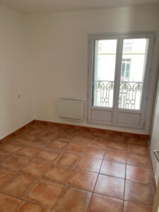Pièce vide lumineuse : murs blancs, carrelage terracotta. Grande fenêtre double avec balustrade fer forgé, radiateur mural.